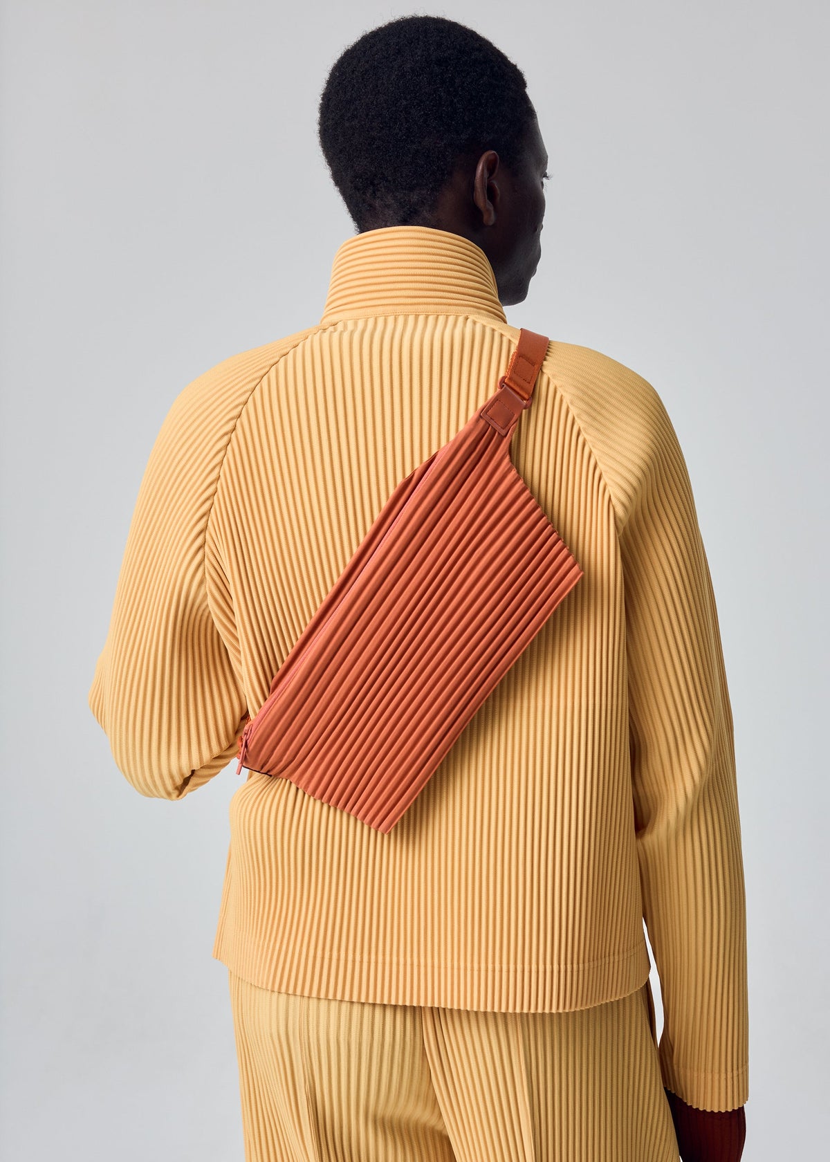 A model wears the HOMME PLISSÉ ISSEY MIYAKE PLEATS SLIM BAG pleats-slim-bag-bag-shade-vermillion-1.